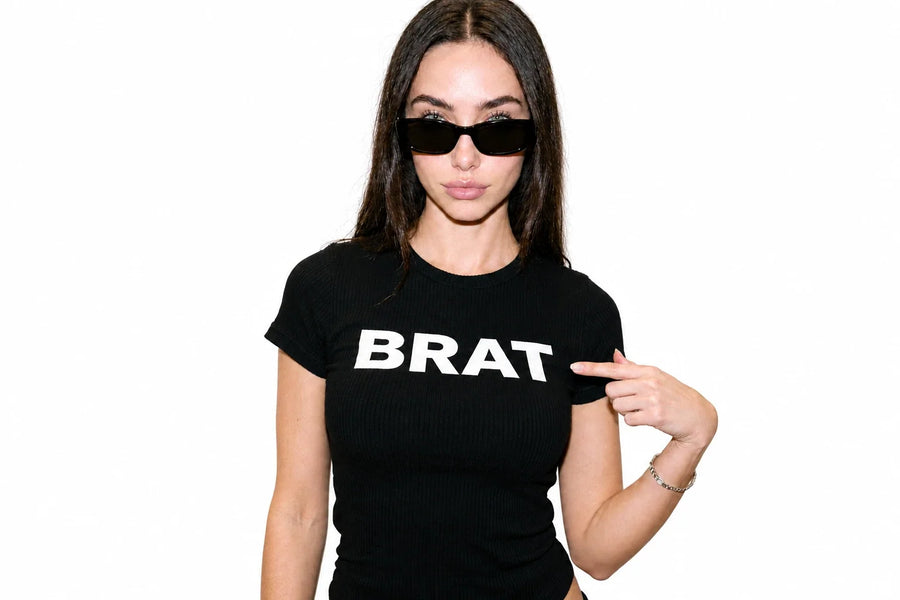 Une femme portant des lunettes noires pointe le mot BRAT inscrit en blanc sur son t-shirt noir.